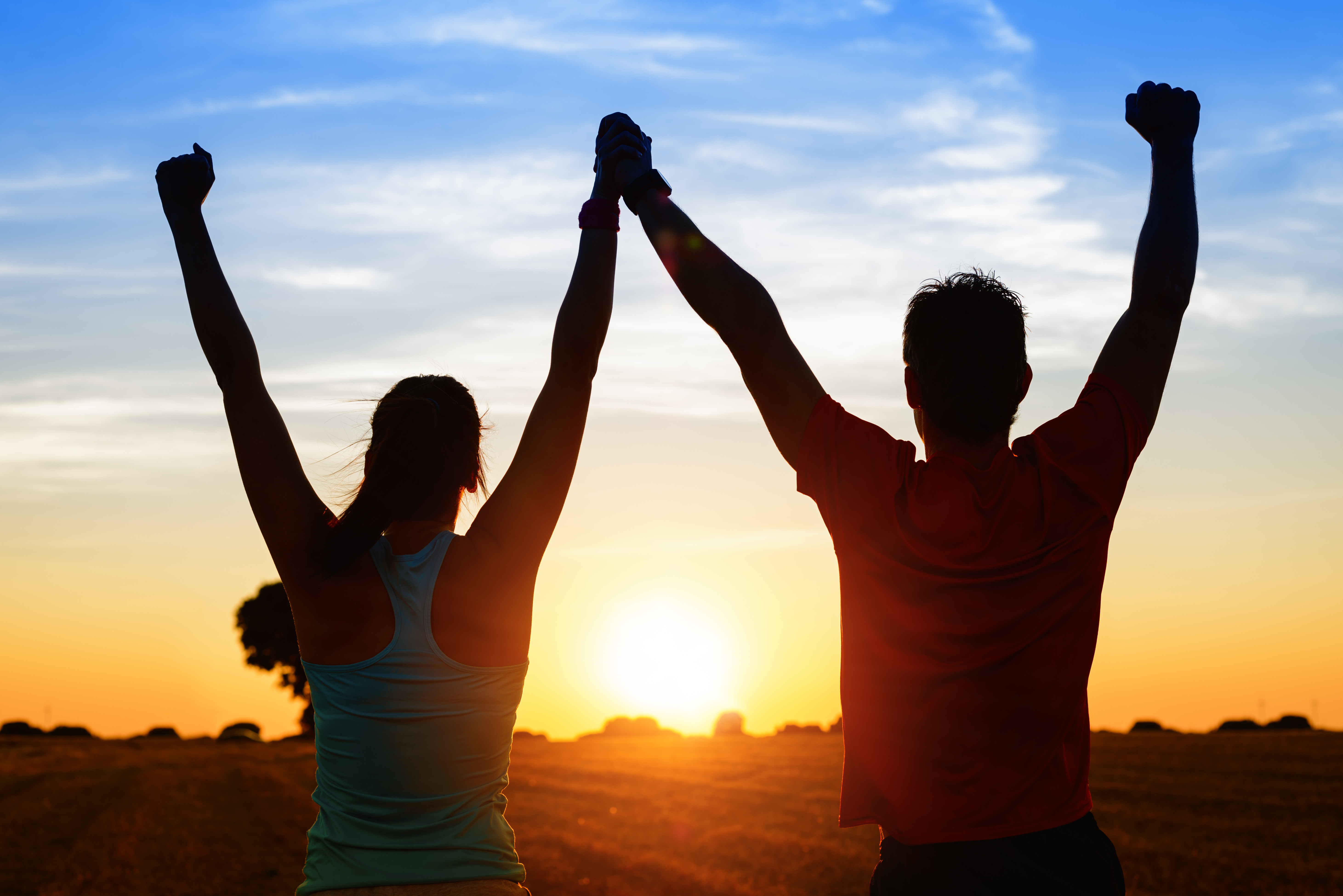 Successful couple of young athletes raising arms to golden summer sunset sky after training. Fitness man and woman with arms up celebrating sport goals after exercising in countryside field.